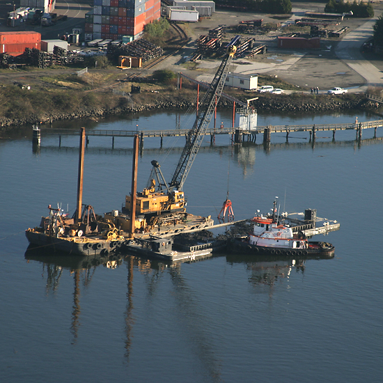 Aerial photo of a yellow excavator crane mounted on a barge near the Duwamish Diagonal. A white tugboat is supporting. In the background is the shoreline with industry and shipping containers.