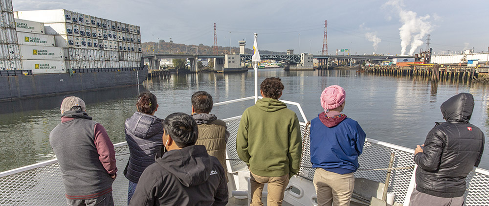 Photo of 7 community members sailing the Duwamish during Port of Seattle's 'Port 101' tour. The people are standing at the front of a white boat, which is approaching the South Park Bridge from the south.