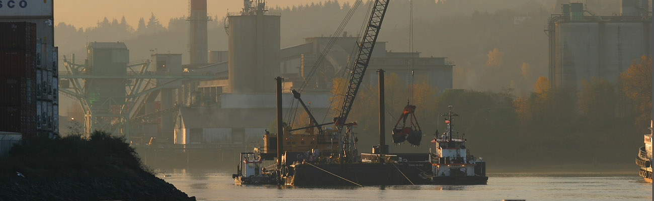 Photo of barge on waterway being loaded with sediment with excavator. Two tugboats are supporting the work.