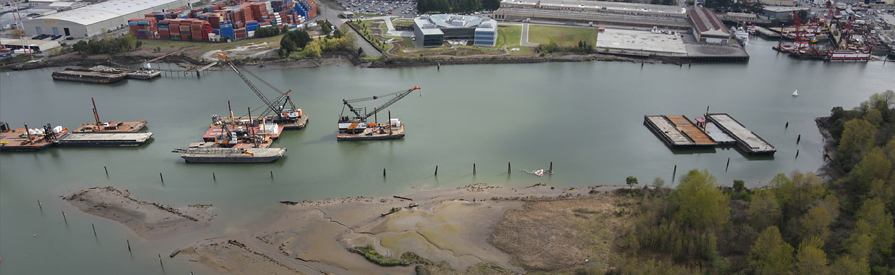 Aerial view of multiple barges traveling south on the Lower Duwamish Waterway. In the background are shipping containers and industrial buildings.