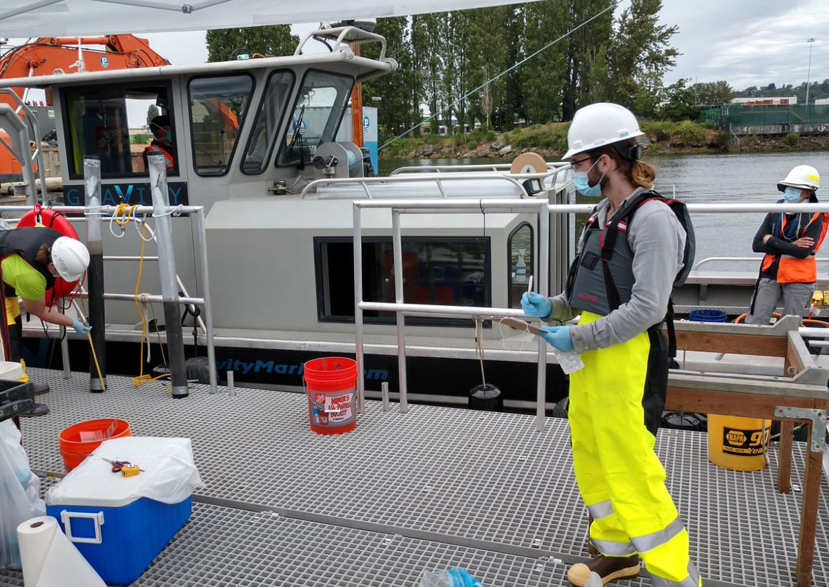 Photo of sampling crew on two boats wearing protective gear. On the boat deck are buckets and a cooler. 