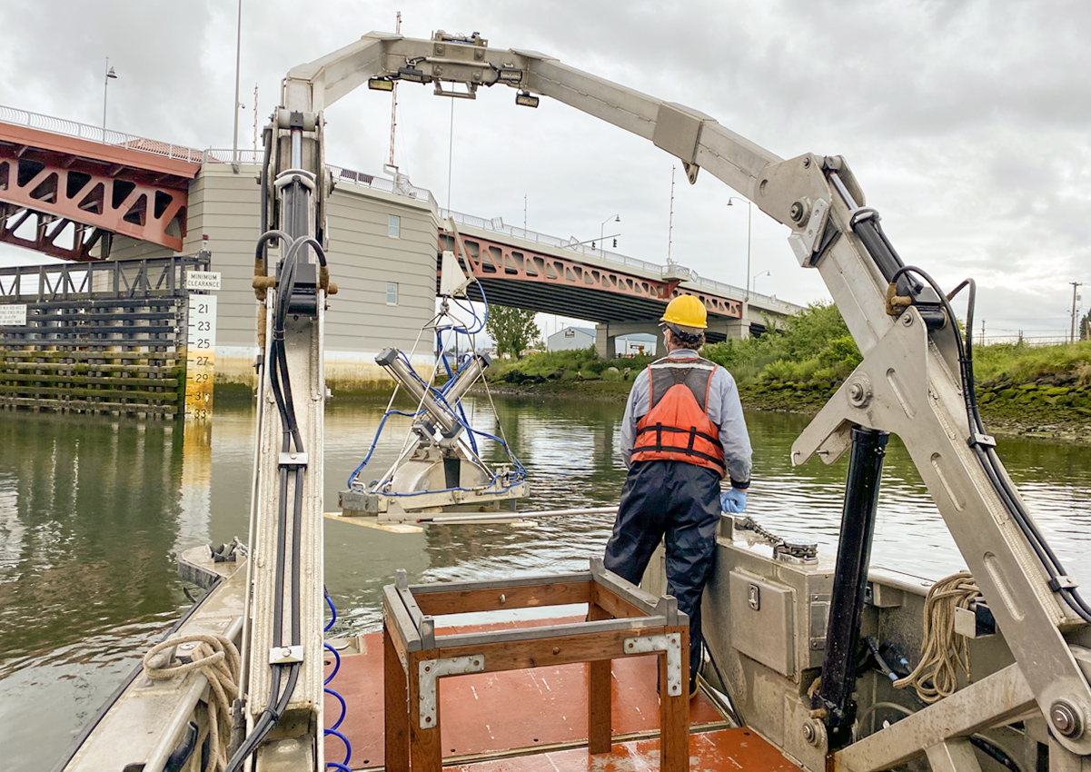 Photo of crews collecting a “grab sample” from the waterway. This image shows a man in safety gear on a boat with equipment, and the South Park Bridge in the distance.