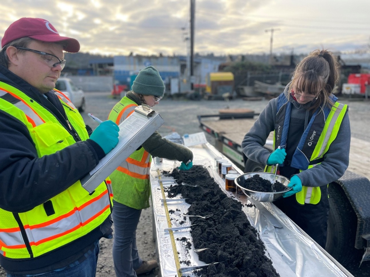 Geosyntec Dec 27 2025 Photo of three people wearing safety vests. The people are looking at sediment core samples on a table. They are outside.